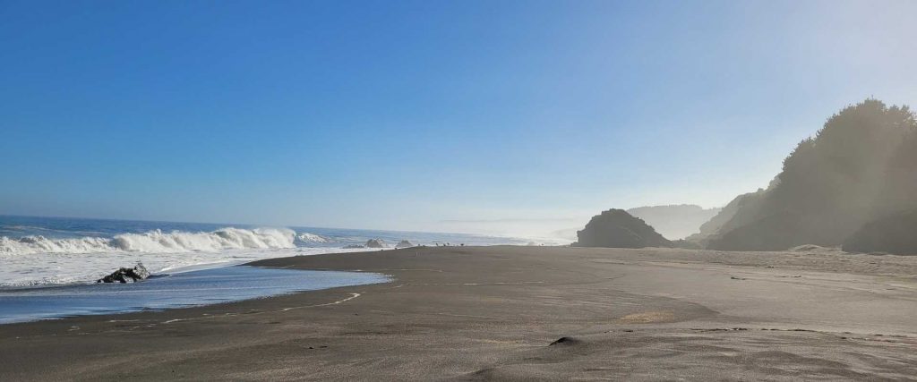 Pazifikküste mit Wellen, dunklem Strand, Felsen und blauem Himmel